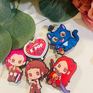 Pu&ograve; includere: Una collezione di ciondoli colorati a tema K-Pop. I ciondoli includono un gatto blu con un cappello, diverse figure in stile anime e un ciondolo a forma di cuore con la scritta "I love K-Pop".