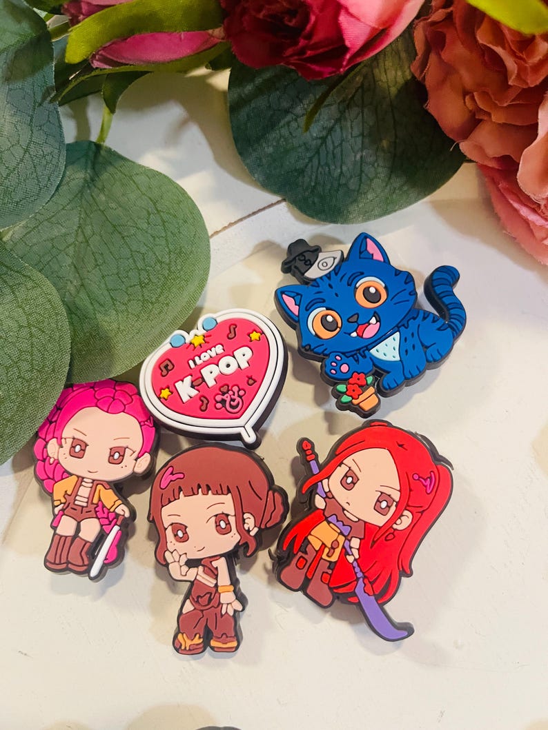 Pu&ograve; includere: Una collezione di ciondoli colorati a tema K-Pop. I ciondoli includono un gatto blu con un cappello, diverse figure in stile anime e un ciondolo a forma di cuore con la scritta "I love K-Pop".