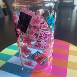 Op de afbeelding: Een doorzichtige plastic beker met een houten deksel en een rietje. De beker is versierd met roze kersenbloesem stickers en boekomslagen met de tekst "It Ends With Us" en "Heart Bones" van Colleen Hoover.
