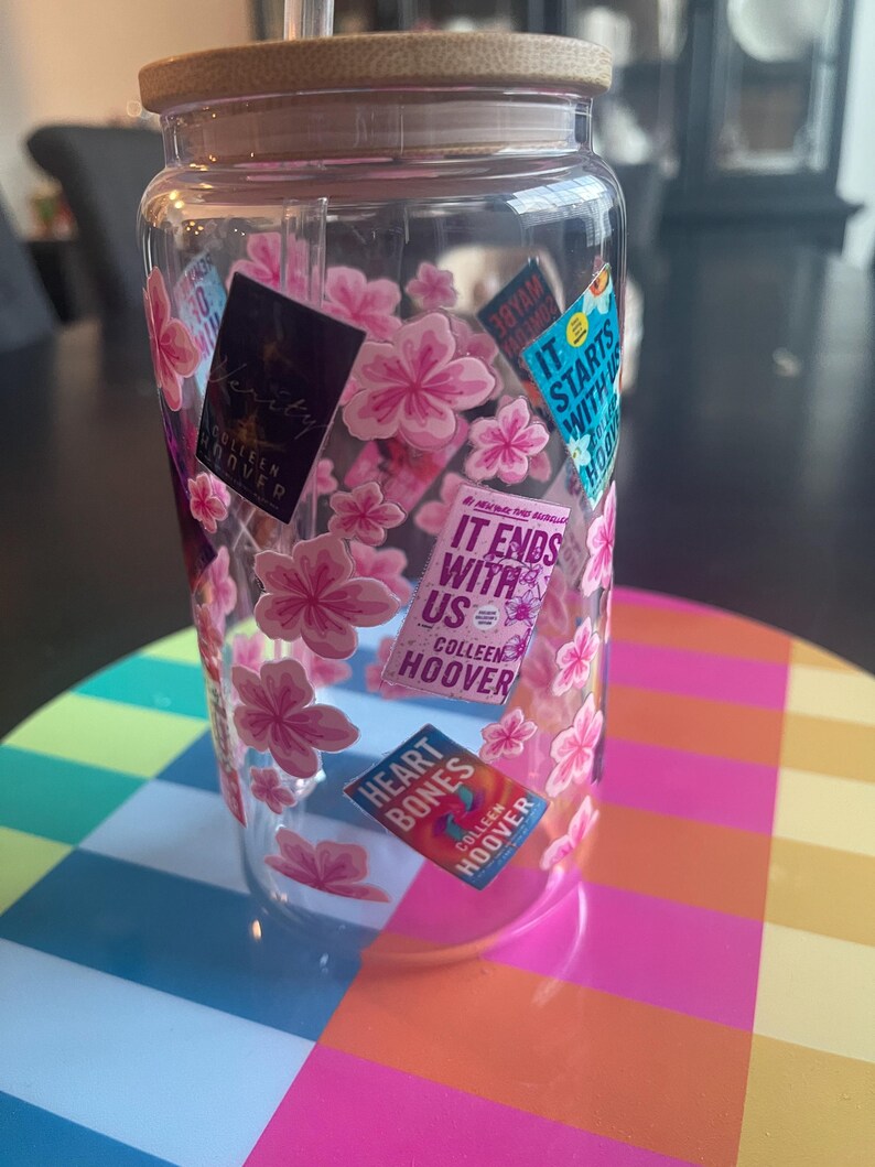 Op de afbeelding: Een doorzichtige plastic beker met een houten deksel en een rietje. De beker is versierd met roze kersenbloesem stickers en boekomslagen met de tekst "It Ends With Us" en "Heart Bones" van Colleen Hoover.