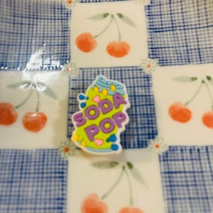 Pu&ograve; includere: Un piatto decorativo con un motivo a quadri blu e bianchi e illustrazioni di ciliegie. Un ciondolo colorato "Soda Pop" &egrave; al centro. Il piatto ha un design stravagante e giocoso.