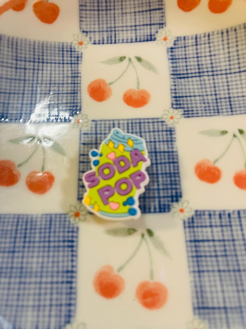 Pu&ograve; includere: Un piatto decorativo con un motivo a quadri blu e bianchi e illustrazioni di ciliegie. Un ciondolo colorato "Soda Pop" &egrave; al centro. Il piatto ha un design stravagante e giocoso.