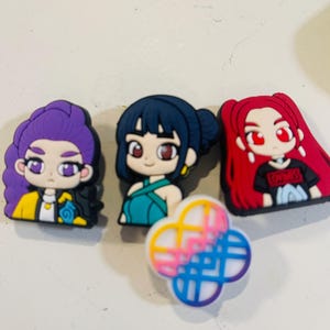 Pu&ograve; includere: Una collezione di ciondoli per scarpe con personaggi dei cartoni animati colorati. I ciondoli presentano tre ragazze in stile anime con vari colori di capelli e abiti, insieme a un design a quadrifoglio in rosa, blu e giallo.