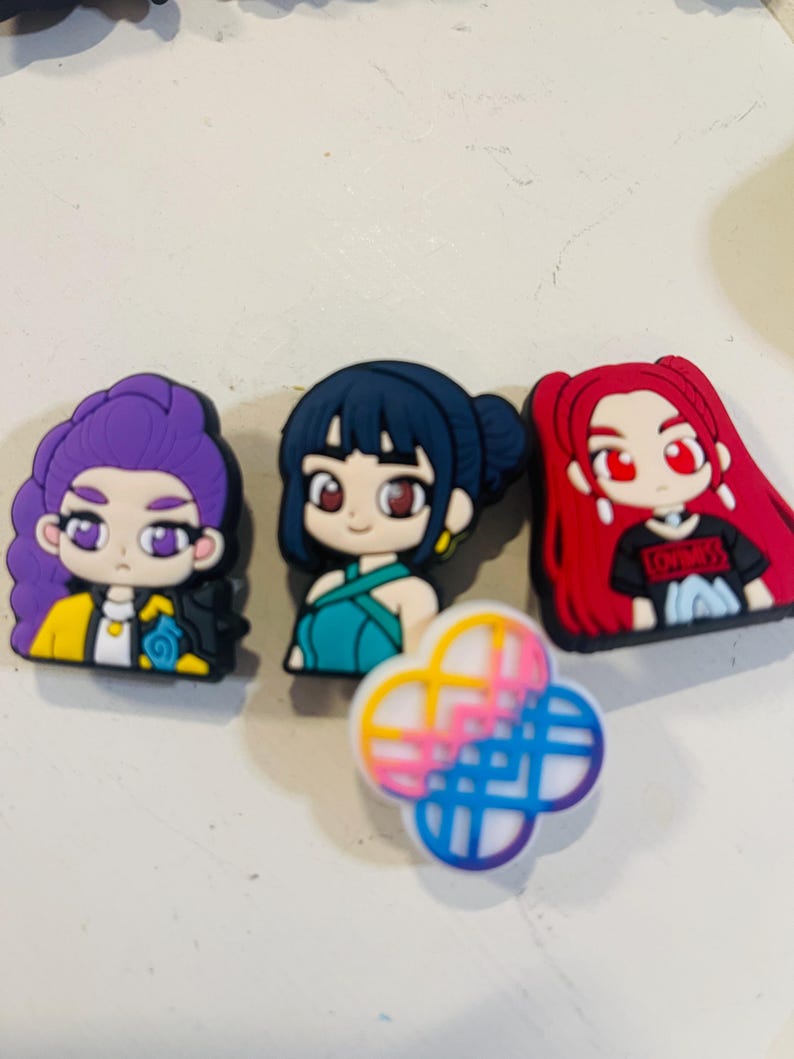Pu&ograve; includere: Una collezione di ciondoli per scarpe con personaggi dei cartoni animati colorati. I ciondoli presentano tre ragazze in stile anime con vari colori di capelli e abiti, insieme a un design a quadrifoglio in rosa, blu e giallo.