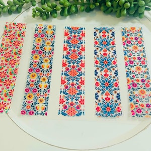 Könnte beinhalten: Fünf dekorative Streifen mit floralen Mustern in verschiedenen Farben. Die Streifen zeigen Blumen in Rosa, Orange, Gelb und Blau auf weißem Hintergrund. Sie sind auf einer weißen Oberfläche mit einer grünen Pflanze im Hintergrund angeordnet.