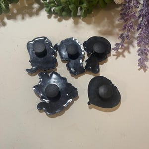 Pu&ograve; includere: Una collezione di sei ciondoli per scarpe grigio scuro con varie forme, tra cui un cuore e delle silhouette. Ogni ciondolo ha un bottone nero e rotondo sul retro per l'attacco. I ciondoli sono disposti su una superficie chiara.
