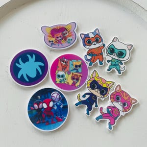 May include: A set of nine colorful iron-on patches featuring cartoon characters. The patches include a spider, a group of cats, and a group of superheroes.