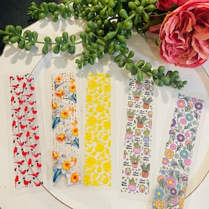 Könnte beinhalten: Set aus fünf dekorativen Lesezeichen mit verschiedenen Designs. Eines zeigt rote Vögel, ein anderes Narzissen, ein drittes gelbe Rosen, ein viertes Topfpflanzen mit dem Text "Plant Mama" und das letzte bunte Blumen. Die Lesezeichen liegen auf einer weißen Oberfläche.