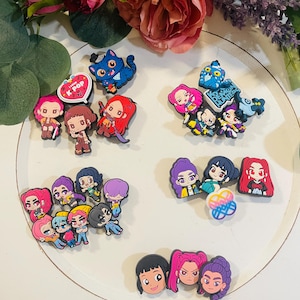 Pu&ograve; includere: Una collezione di ciondoli colorati di personaggi dei cartoni animati, tra cui figure di gatti e umane, disposti su una superficie bianca. Alcuni ciondoli hanno il testo "I love K-POP" e "Demon Hunters". I ciondoli sono in vari colori, tra cui rosa, blu e viola.
