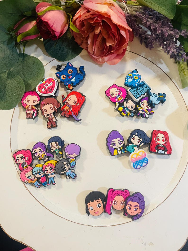 Pu&ograve; includere: Una collezione di ciondoli colorati di personaggi dei cartoni animati, tra cui figure di gatti e umane, disposti su una superficie bianca. Alcuni ciondoli hanno il testo "I love K-POP" e "Demon Hunters". I ciondoli sono in vari colori, tra cui rosa, blu e viola.