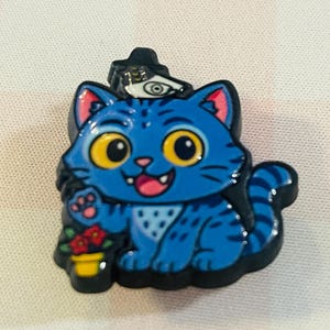 Pu&ograve; includere: Spilla smaltata a forma di gatto blu con contorno nero. Il gatto indossa un cappello con un simbolo oculare e tiene un vaso di fiori rossi. Il gatto ha occhi gialli, lingua rosa e un motivo bianco sulla pancia.