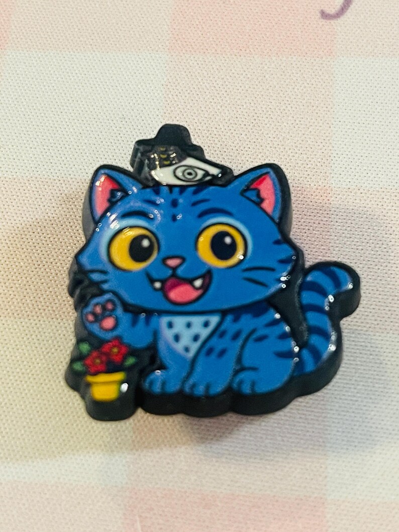 Pu&ograve; includere: Spilla smaltata a forma di gatto blu con contorno nero. Il gatto indossa un cappello con un simbolo oculare e tiene un vaso di fiori rossi. Il gatto ha occhi gialli, lingua rosa e un motivo bianco sulla pancia.