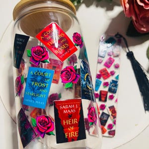 Op de afbeelding: Een doorzichtige glazen beker met een houten deksel en een rietje. De beker is versierd met kleurrijke stickers met boekomslagen uit de serie "A Court of Thorns and Roses" van Sarah J. Maas. De stickers bevatten rozen en de titels "A Court of Thorns and Roses", "A Court of Frost and Starlight" en "Heir of Fire".