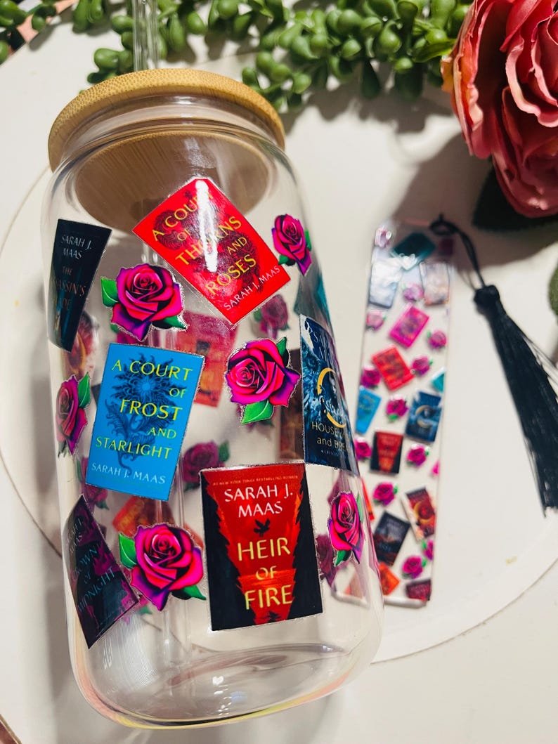 Op de afbeelding: Een doorzichtige glazen beker met een houten deksel en een rietje. De beker is versierd met kleurrijke stickers met boekomslagen uit de serie "A Court of Thorns and Roses" van Sarah J. Maas. De stickers bevatten rozen en de titels "A Court of Thorns and Roses", "A Court of Frost and Starlight" en "Heir of Fire".