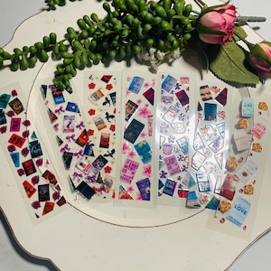 Puede incluir: Un conjunto de seis marcadores de libros con temática de libros con varias portadas y títulos de libros. Los marcadores están en un plato blanco con una planta verde y una rosa rosa.