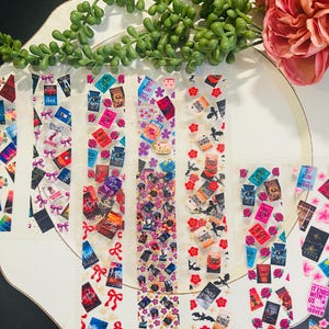 Op de afbeelding: Transparante plakbandstrips versierd met miniatuur boekomslagen, strikken, bloemen en tekst. De tekst bevat "CRASH", "OF HIM", "ONYX STORM" en "IT ENDS WITH US". De strips zijn gerangschikt op een wit oppervlak met groene en roze bloemaccenten.