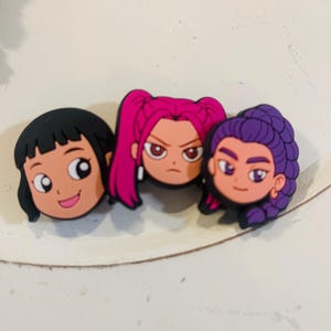 Pu&ograve; includere: Tre ciondoli per scarpe con personaggi dei cartoni animati. Il primo ha i capelli neri e un'espressione sorridente. Il secondo ha i capelli rosa e un'espressione arrabbiata. Il terzo ha i capelli viola e un'espressione neutra.