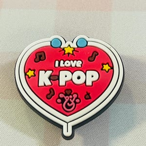 Pu&ograve; includere: Un ciondolo a forma di cuore con la scritta "I LOVE K-POP" in lettere bianche su sfondo rosso. Il ciondolo ha un bordo bianco ed &egrave; decorato con note musicali, stelle e cerchi blu. Probabilmente &egrave; un accessorio per scarpe.