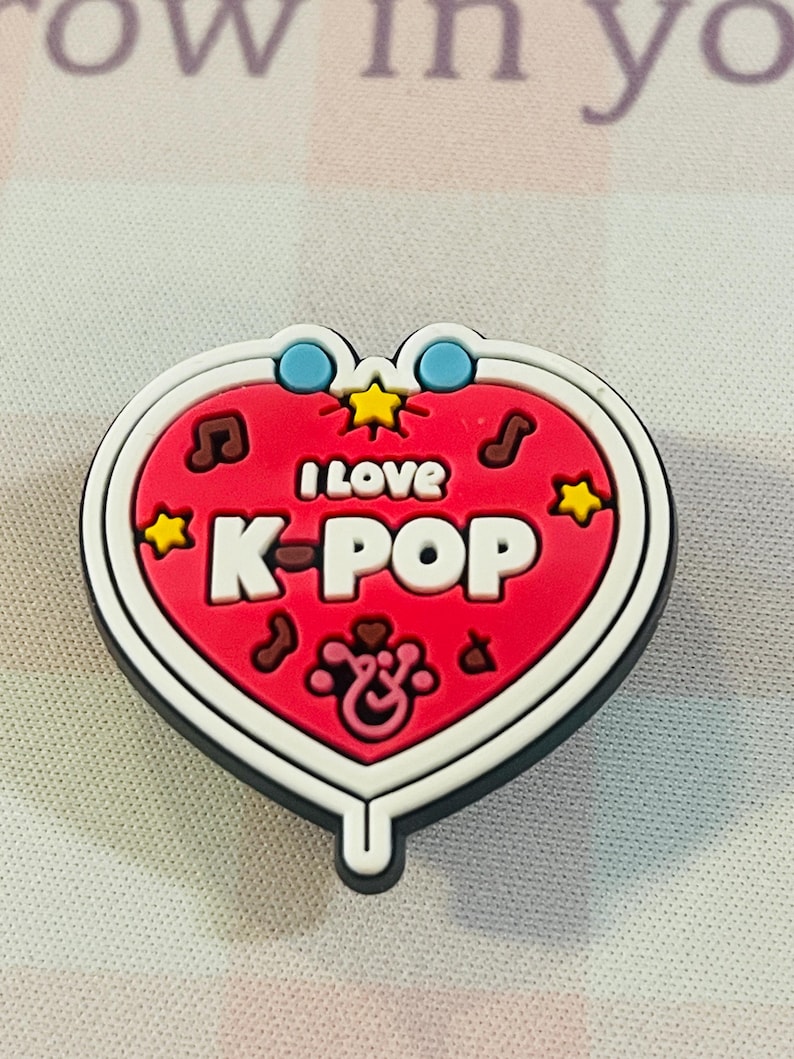 Pu&ograve; includere: Un ciondolo a forma di cuore con la scritta "I LOVE K-POP" in lettere bianche su sfondo rosso. Il ciondolo ha un bordo bianco ed &egrave; decorato con note musicali, stelle e cerchi blu. Probabilmente &egrave; un accessorio per scarpe.