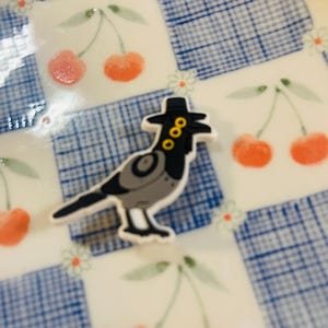Pu&ograve; includere: Un uccello stravagante in stile cartone animato con un cappello nero con accenti dorati. L'uccello &egrave; grigio e nero con un contorno bianco. Lo sfondo &egrave; una superficie bianca con un motivo a scacchi blu e illustrazioni di ciliegie.