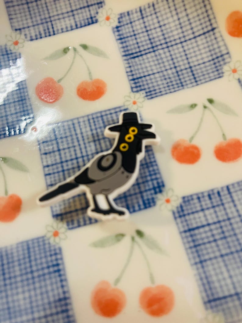 Pu&ograve; includere: Un uccello stravagante in stile cartone animato con un cappello nero con accenti dorati. L'uccello &egrave; grigio e nero con un contorno bianco. Lo sfondo &egrave; una superficie bianca con un motivo a scacchi blu e illustrazioni di ciliegie.