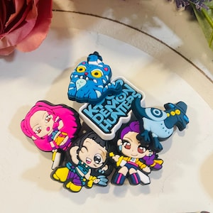 Pu&ograve; includere: Una collezione di ciondoli per scarpe con personaggi dei cartoni animati colorati. I ciondoli presentano vari personaggi con capelli e abiti vivaci, insieme al testo "K Demon Hunters". I ciondoli sono realizzati in materiale gommoso.