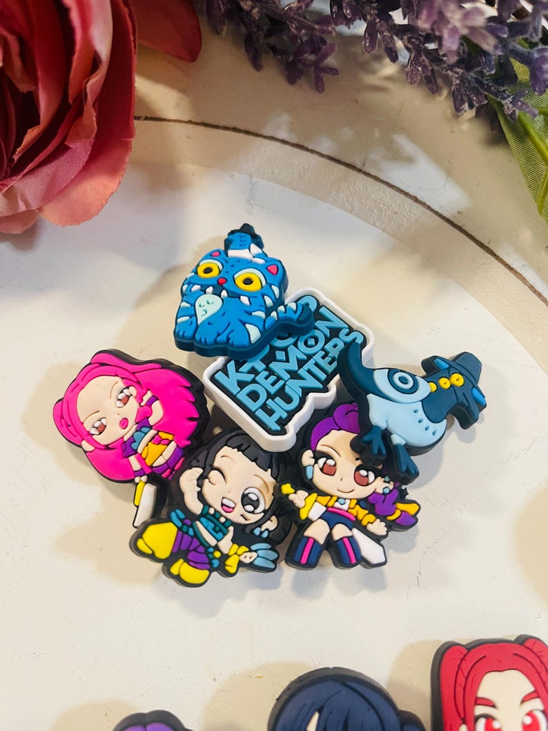 Pu&ograve; includere: Una collezione di ciondoli per scarpe con personaggi dei cartoni animati colorati. I ciondoli presentano vari personaggi con capelli e abiti vivaci, insieme al testo "K Demon Hunters". I ciondoli sono realizzati in materiale gommoso.