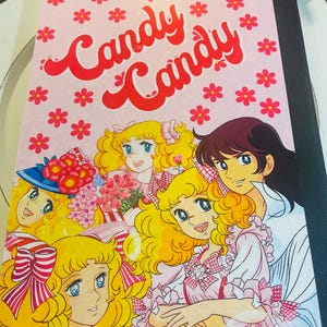 May include: A notebook featuring the title "Candy Candy" in red script on a pink background, adorned with red flower illustrations. The cover showcases a colorful anime-style illustration of several characters with blonde hair and blue eyes.