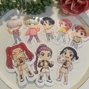 Peut inclure: Une collection d'autocollants holographiques représentant des illustrations de personnages de style anime. Les autocollants présentent diverses figures en tenues colorées, certaines portant des vestes de style militaire et d'autres des tenues décontractées. Les autocollants sont sur une surface blanche.