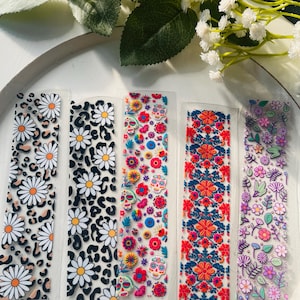 Puede incluir: Cinco hojas de pegatinas transparentes con diferentes diseños florales y de calaveras. Las pegatinas están sobre un fondo blanco.