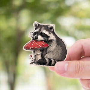 May include: A raccoon sticker with a red and white mushroom. The raccoon is black, white, and gray, and is holding the mushroom. The sticker is held up by a hand against a blurred green background.