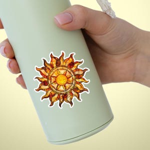 May include: A light green insulated water bottle with a decorative sun sticker. The sun design features a yellow center with orange and brown rays. The bottle has a white rope handle.