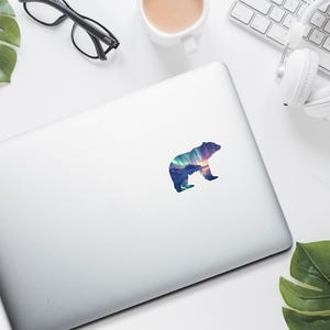 May include: A colourful bear sticker with a landscape design, featuring shades of blue, purple, and orange. The sticker is on a silver laptop, surrounded by office supplies, including glasses, headphones, and a coffee cup.