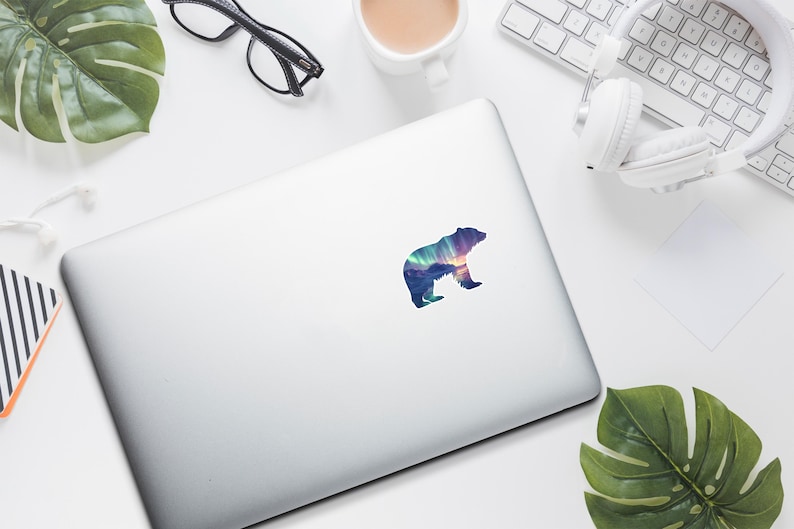 May include: A colourful bear sticker with a landscape design, featuring shades of blue, purple, and orange. The sticker is on a silver laptop, surrounded by office supplies, including glasses, headphones, and a coffee cup.