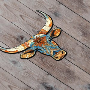 Puede incluir: Recorte decorativo de madera de una cabeza de toro con cuernos largos. La cabeza del toro está decorada con un colorido patrón floral y geométrico en tonos naranja, azul y amarillo. El recorte se coloca sobre una superficie de madera.