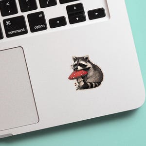 May include: A sticker featuring a detailed illustration of a raccoon holding a red and white mushroom. The raccoon is depicted in shades of gray and black, with a white face and striped tail. The sticker is placed on a white laptop.