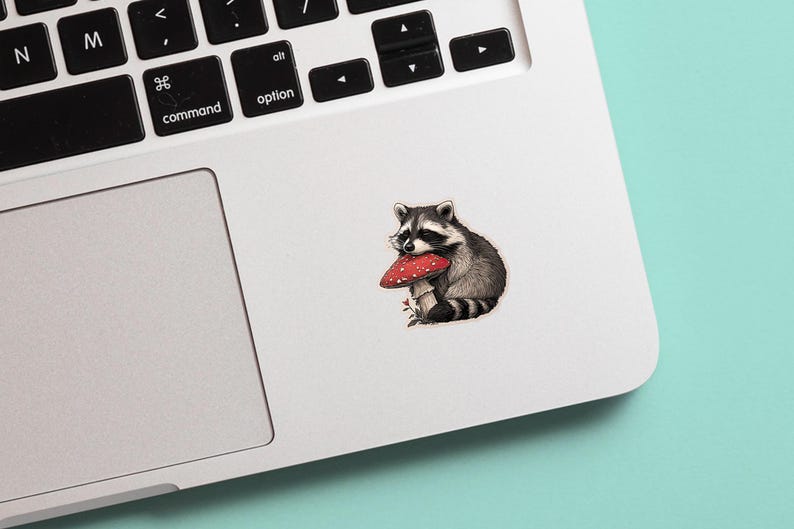May include: A sticker featuring a detailed illustration of a raccoon holding a red and white mushroom. The raccoon is depicted in shades of gray and black, with a white face and striped tail. The sticker is placed on a white laptop.