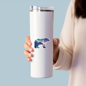 May include: A white stainless steel travel mug with a bear sticker. The sticker shows a bear silhouette filled with a landscape of mountains, aurora borealis, and a sunset. The mug is held by a person.