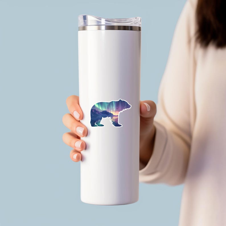 May include: A white stainless steel travel mug with a bear sticker. The sticker shows a bear silhouette filled with a landscape of mountains, aurora borealis, and a sunset. The mug is held by a person.