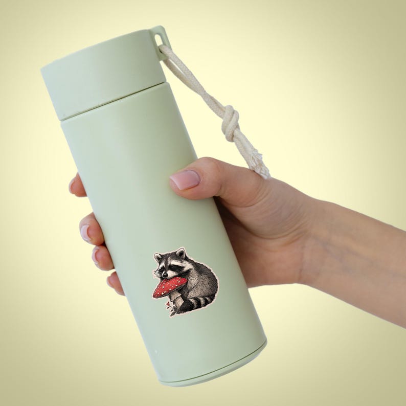 May include: A light green insulated water bottle with a white rope handle. A sticker of a raccoon holding a red mushroom is on the bottle. The bottle is held by a person.