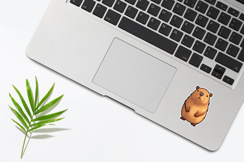 Pode incluir: Um adesivo de capivara de desenho animado marrom e branco em um laptop prateado.