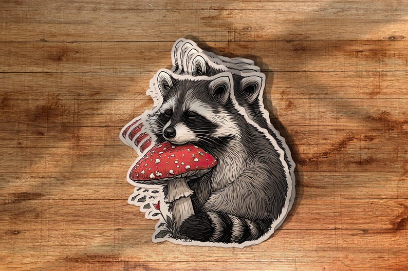 May include: A sticker featuring a detailed illustration of a raccoon hugging a red mushroom with white spots. The raccoon is in shades of gray and black, with a white face and a striped tail. The sticker is on a wooden surface.