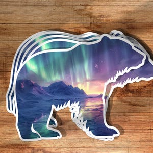 May include: A layered sticker in the shape of a bear, featuring a landscape scene. The scene inside the bear silhouette depicts mountains, a body of water, and a colourful aurora borealis in shades of green, purple, and blue.