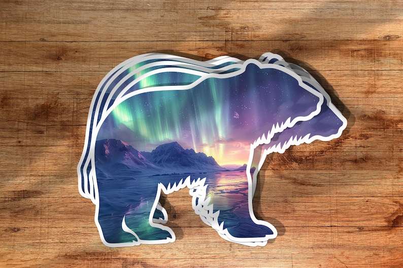 May include: A layered sticker in the shape of a bear, featuring a landscape scene. The scene inside the bear silhouette depicts mountains, a body of water, and a colourful aurora borealis in shades of green, purple, and blue.