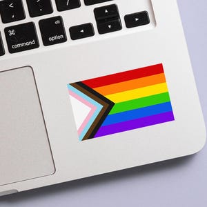 May include: A rectangular sticker featuring the Progress Pride flag, with the traditional rainbow stripes and additional stripes representing transgender, Black, and brown communities. The sticker is placed on a silver laptop.