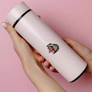 May include: A light pink insulated water bottle with a black top and bottom rim. A raccoon sticker with a red mushroom is on the side. The bottle is held by two hands against a pink background.