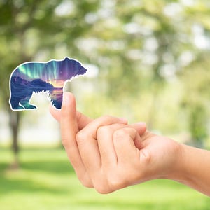 May include: A bear-shaped sticker with a landscape design depicting mountains, water, and the aurora borealis. The sticker is held up against a blurred green background.