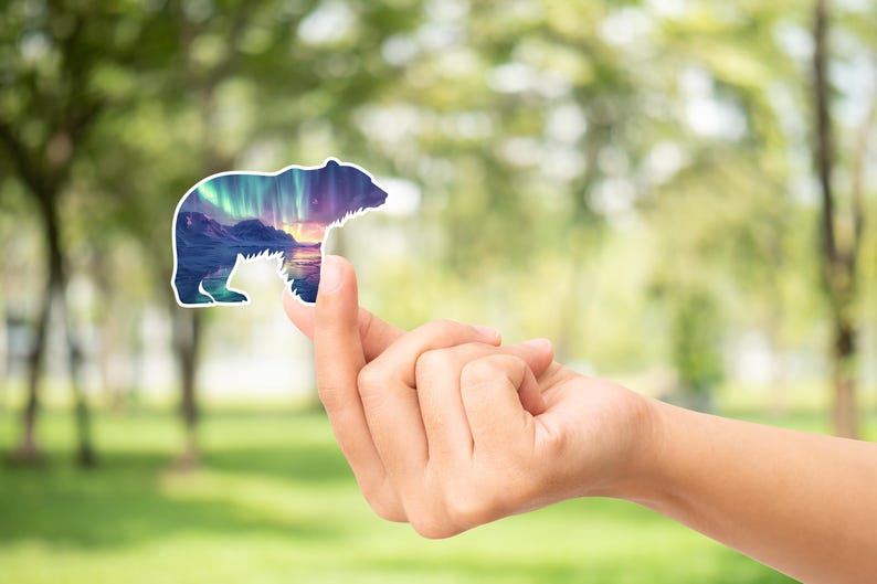 May include: A bear-shaped sticker with a landscape design depicting mountains, water, and the aurora borealis. The sticker is held up against a blurred green background.