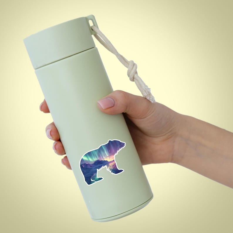 May include: A pale green insulated water bottle with a white rope handle. A sticker of a bear silhouette with a colourful aurora borealis design is affixed to the bottle. The bottle is held by a person.