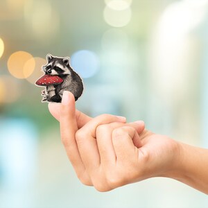 May include: A raccoon sticker with a red and white mushroom. The raccoon is black, gray, and white. The sticker is held between two fingers. The background is blurred with bokeh lights.